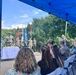 A military memorial or commemorative ceremony is taking place outdoors under a blue canopy tent. A color guard is presenting multiple flags including the American flag and what appears to be the South Korean flag, suggesting a joint US-Korean military event. Attendees including both military personnel in camouflage uniforms and civilians are seated facing a stage with a white-draped table adorned with floral wreaths.