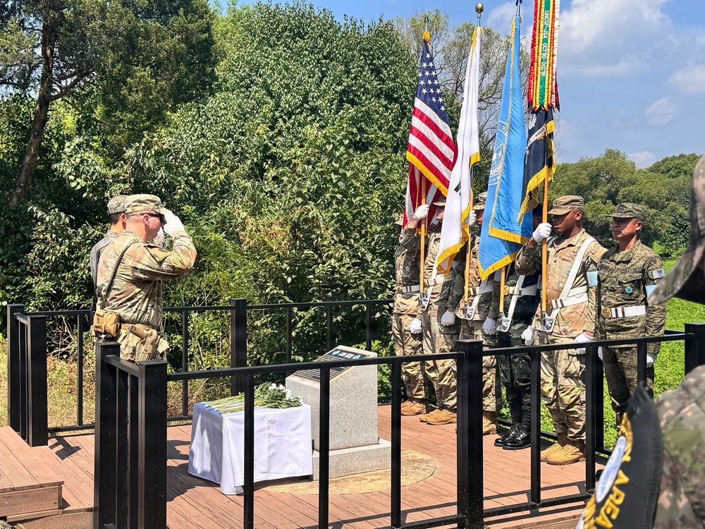 A military ceremony is taking place on an outdoor wooden deck platform, featuring a color guard presenting multiple flags including the United States flag and a United Nations flag. A soldier on the left is rendering a salute toward the color guard, while personnel in mixed military uniforms stand at attention. A draped table with flowers is visible, suggesting a memorial or commemorative event.