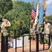 A military ceremony is taking place on an outdoor wooden deck platform, featuring a color guard presenting multiple flags including the United States flag and a United Nations flag. A soldier on the left is rendering a salute toward the color guard, while personnel in mixed military uniforms stand at attention. A draped table with flowers is visible, suggesting a memorial or commemorative event.