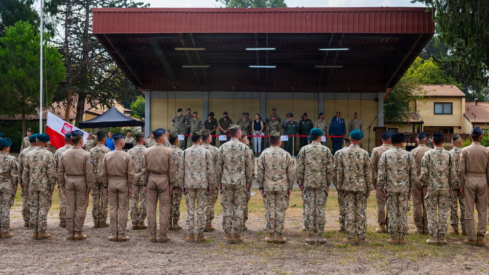 A military formation ceremony is taking place outdoors at what appears to be a military installation. Rows of soldiers in various camouflage uniforms stand at attention facing a covered stage where officials and dignitaries are assembled. A Polish national flag (white and red with eagle emblem) is visible on the left side of the formation.