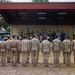 A military formation ceremony is taking place outdoors at what appears to be a military installation. Rows of soldiers in various camouflage uniforms stand at attention facing a covered stage where officials and dignitaries are assembled. A Polish national flag (white and red with eagle emblem) is visible on the left side of the formation.