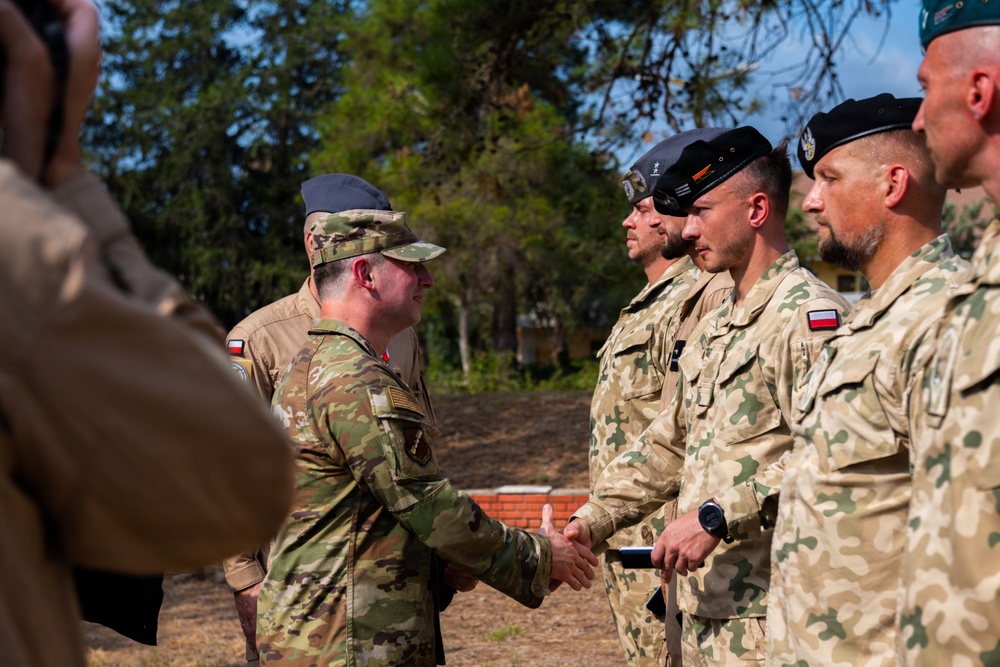 A U.S. Army soldier in OCP (Operational Camouflage Pattern) uniform is shaking hands with a soldier wearing Polish military camouflage uniform, suggesting a formal military greeting or ceremony. Several other Polish military personnel are standing in a line, wearing distinctive tan/green camouflage uniforms and black or green berets. The setting appears to be an outdoor military installation or training area.