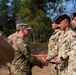 A U.S. Army soldier in OCP (Operational Camouflage Pattern) uniform is shaking hands with a soldier wearing Polish military camouflage uniform, suggesting a formal military greeting or ceremony. Several other Polish military personnel are standing in a line, wearing distinctive tan/green camouflage uniforms and black or green berets. The setting appears to be an outdoor military installation or training area.