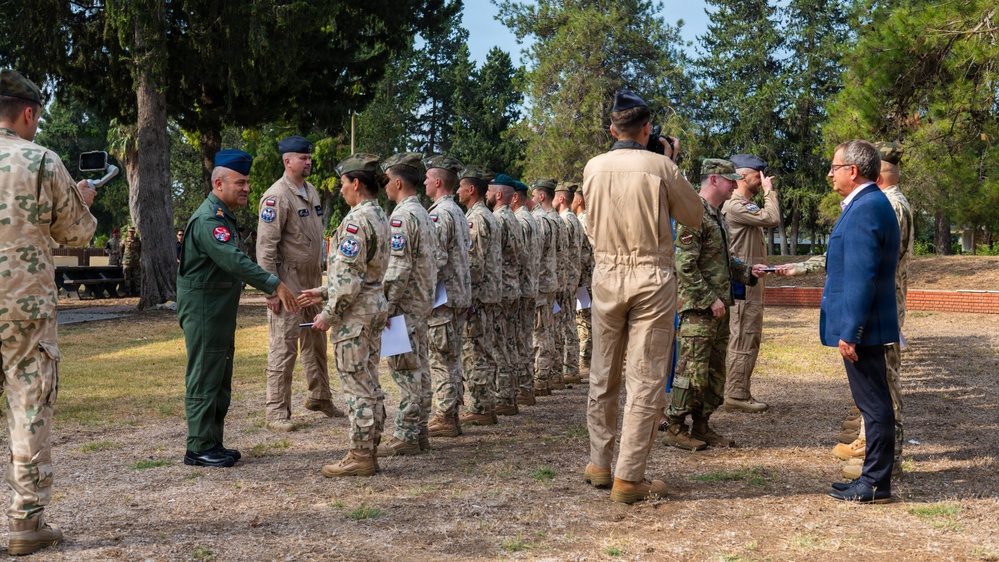 A military ceremony is taking place outdoors in a wooded area, where a senior officer in a green flight suit is greeting and shaking hands with a line of multinational military personnel in various uniforms. A civilian in a blue suit observes from the right side. The event appears to be a formal recognition or award ceremony involving multiple nations' armed forces.