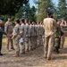 A military ceremony is taking place outdoors in a wooded area, where a senior officer in a green flight suit is greeting and shaking hands with a line of multinational military personnel in various uniforms. A civilian in a blue suit observes from the right side. The event appears to be a formal recognition or award ceremony involving multiple nations' armed forces.