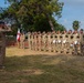 A military ceremony is taking place outdoors on a grassy field, with a large formation of soldiers in various camouflage uniforms rendering a salute. Two officers in tan/khaki uniforms and blue berets face the formation and return the salute. A white and red flag, consistent with the Polish national flag, is visible in the background near the formation.