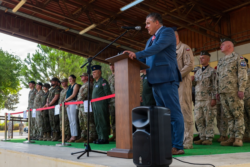 A civilian official in a blue suit is delivering a speech at a podium during a multinational military ceremony or event. Multiple military personnel in various camouflage uniforms are standing in formation behind him on a covered outdoor stage. The presence of different uniform patterns and national patches suggests a NATO or coalition military gathering.