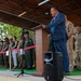 A civilian official in a blue suit is delivering a speech at a podium during a multinational military ceremony or event. Multiple military personnel in various camouflage uniforms are standing in formation behind him on a covered outdoor stage. The presence of different uniform patterns and national patches suggests a NATO or coalition military gathering.
