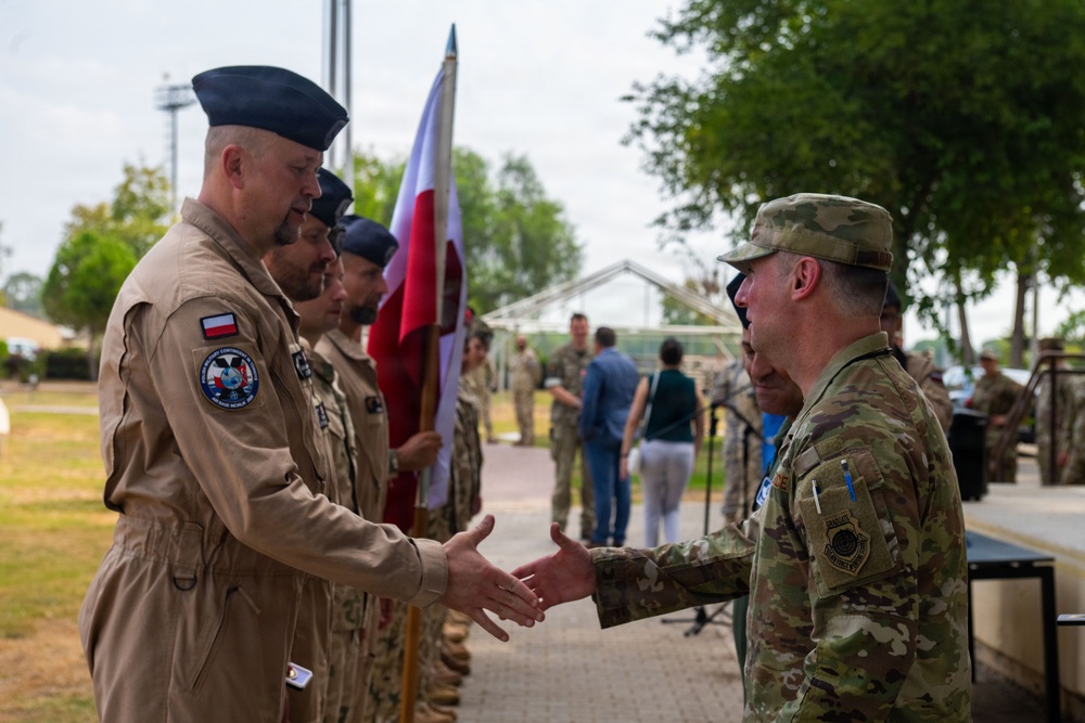 A military ceremony appears to be taking place outdoors, showing a handshake between a Polish Air Force officer in tan/khaki flight suit and a U.S. military personnel in OCP (Operational Camouflage Pattern) uniform. A Polish flag is visible being carried by personnel in the background, suggesting a formal bilateral military event such as a transfer of authority or welcome ceremony. Multiple military personnel from what appear to be Polish and U.S. forces are present in formation.