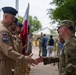 A military ceremony appears to be taking place outdoors, showing a handshake between a Polish Air Force officer in tan/khaki flight suit and a U.S. military personnel in OCP (Operational Camouflage Pattern) uniform. A Polish flag is visible being carried by personnel in the background, suggesting a formal bilateral military event such as a transfer of authority or welcome ceremony. Multiple military personnel from what appear to be Polish and U.S. forces are present in formation.