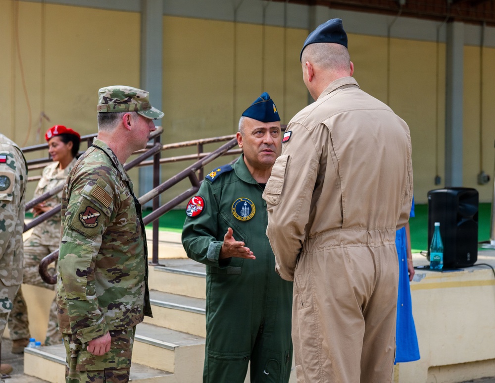The image shows a multinational military gathering where officers from at least two or three different nations are engaged in conversation. A central figure wearing a dark green flight suit with a Turkish flag patch and unit insignia appears to be speaking to colleagues. The setting appears to be an indoor or covered military facility.
