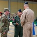 The image shows a multinational military gathering where officers from at least two or three different nations are engaged in conversation. A central figure wearing a dark green flight suit with a Turkish flag patch and unit insignia appears to be speaking to colleagues. The setting appears to be an indoor or covered military facility.