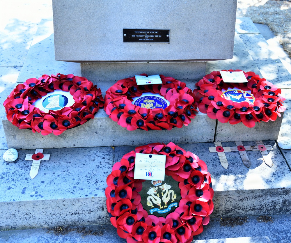 A memorial monument base displays four red poppy wreaths, a traditional British symbol of remembrance for fallen soldiers. A dedication plaque on the stone base reads 'Unveiled on 18th June 2007 by The Viscount Lord Slim OBE DL and Holly Wingate.' The wreaths bear various organizational insignia and handwritten tribute cards, surrounded by small memorial crosses and individual poppies placed on the ground.
