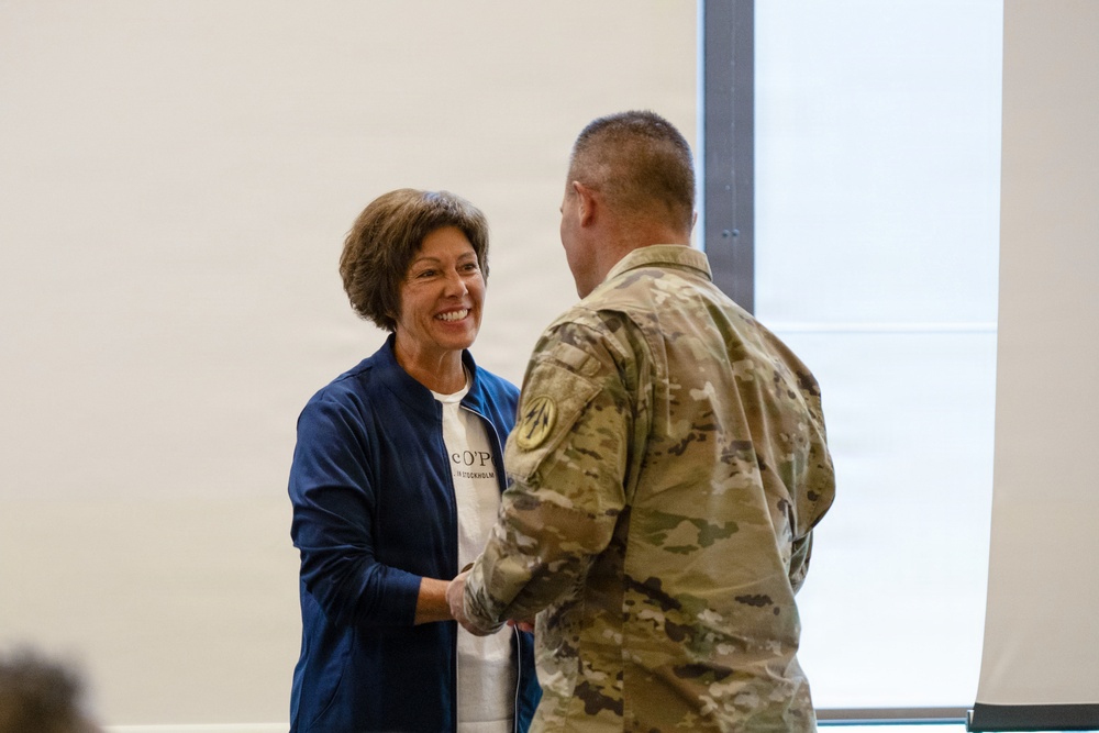 A woman in a blue jacket and white t-shirt is shaking hands with a military service member wearing MultiCam/OCP camouflage uniform. The woman is smiling broadly, suggesting a positive or ceremonial interaction. The setting appears to be an indoor facility with a plain white wall in the background.