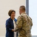 A woman in a blue jacket and white t-shirt is shaking hands with a military service member wearing MultiCam/OCP camouflage uniform. The woman is smiling broadly, suggesting a positive or ceremonial interaction. The setting appears to be an indoor facility with a plain white wall in the background.