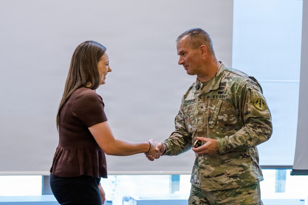 A U.S. Army soldier in OCP (Operational Camouflage Pattern) uniform is shaking hands with a civilian woman in a formal indoor setting. The soldier, identified by his name tape as 'Carpenter,' appears to be presenting or receiving a coin, a common military recognition ceremony. Both individuals are smiling, suggesting a positive ceremonial exchange.