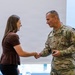 A U.S. Army soldier in OCP (Operational Camouflage Pattern) uniform is shaking hands with a civilian woman in a formal indoor setting. The soldier, identified by his name tape as 'Carpenter,' appears to be presenting or receiving a coin, a common military recognition ceremony. Both individuals are smiling, suggesting a positive ceremonial exchange.