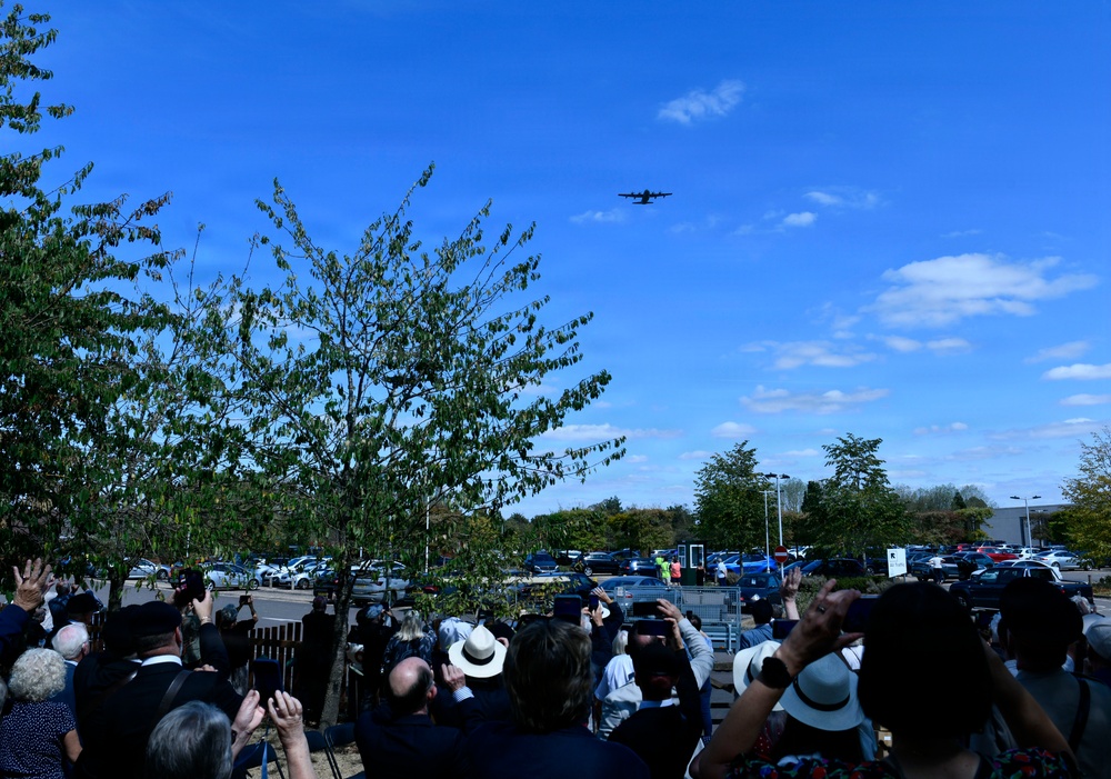 A crowd of people gathered outdoors are looking up and photographing a large military-style aircraft flying overhead in a clear blue sky. The scene appears to be a public event or ceremony, possibly a memorial or airshow, given the formal attire worn by several attendees. The gathering takes place near a car park surrounded by trees in what appears to be a suburban or semi-rural setting.