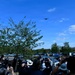 A crowd of people gathered outdoors are looking up and photographing a large military-style aircraft flying overhead in a clear blue sky. The scene appears to be a public event or ceremony, possibly a memorial or airshow, given the formal attire worn by several attendees. The gathering takes place near a car park surrounded by trees in what appears to be a suburban or semi-rural setting.