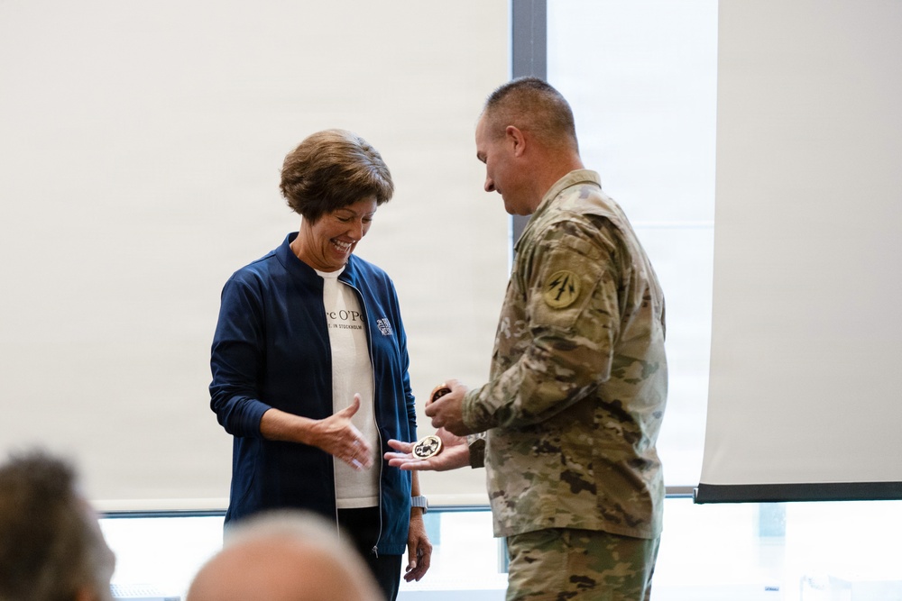 A military service member in camouflage uniform is presenting what appears to be a challenge coin to a civilian woman wearing a navy blue jacket. The woman is smiling and holding out her hand to receive the coin in what appears to be an indoor ceremonial or recognition event. The setting appears to be a modern facility with large windows in the background.