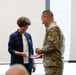 A military service member in camouflage uniform is presenting what appears to be a challenge coin to a civilian woman wearing a navy blue jacket. The woman is smiling and holding out her hand to receive the coin in what appears to be an indoor ceremonial or recognition event. The setting appears to be a modern facility with large windows in the background.