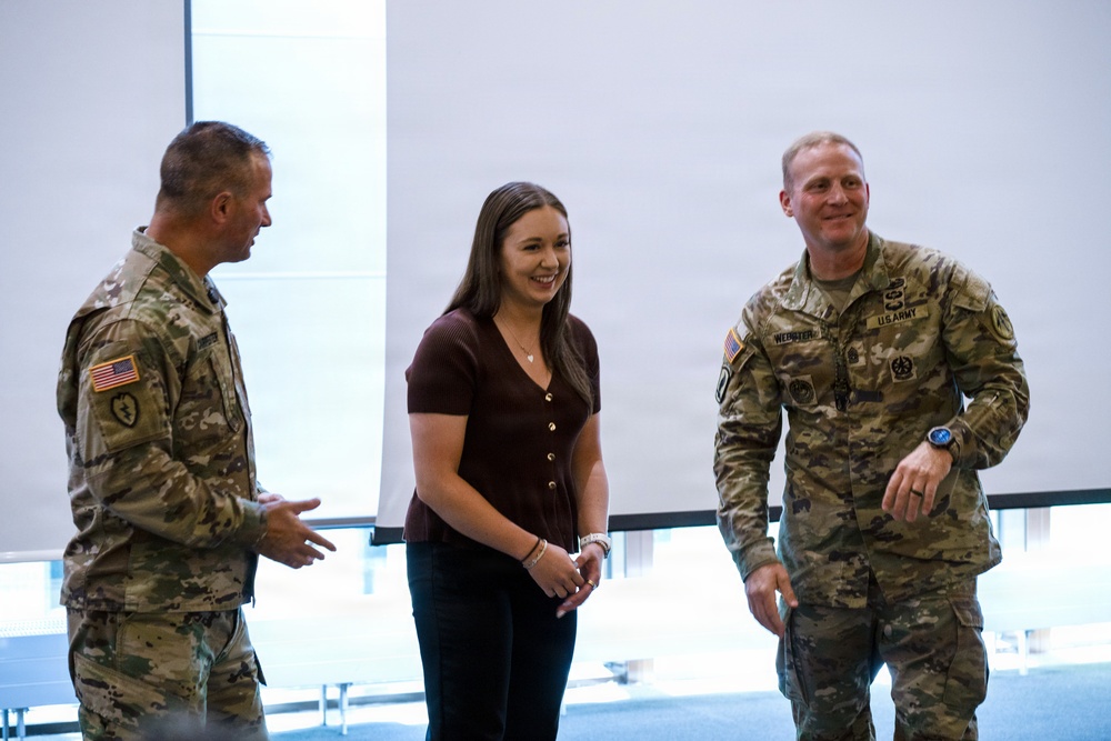 Two U.S. Army soldiers in OCP (Operational Camouflage Pattern) uniforms are engaged in a friendly conversation with a civilian woman in a casual indoor setting. All three individuals appear relaxed and smiling, suggesting a positive social or ceremonial interaction. The setting appears to be a modern facility with large windows in the background.