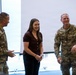 Two U.S. Army soldiers in OCP (Operational Camouflage Pattern) uniforms are engaged in a friendly conversation with a civilian woman in a casual indoor setting. All three individuals appear relaxed and smiling, suggesting a positive social or ceremonial interaction. The setting appears to be a modern facility with large windows in the background.