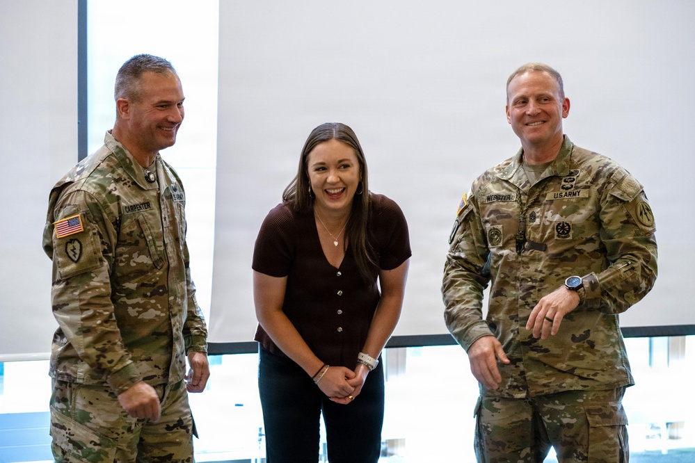 Three individuals are posing together and smiling in what appears to be a modern indoor facility. Two U.S. Army soldiers in OCP (Operational Camouflage Pattern) uniforms flank a civilian woman in casual attire. The atmosphere appears celebratory or ceremonial based on their expressions and body language.