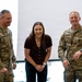 Three individuals are posing together and smiling in what appears to be a modern indoor facility. Two U.S. Army soldiers in OCP (Operational Camouflage Pattern) uniforms flank a civilian woman in casual attire. The atmosphere appears celebratory or ceremonial based on their expressions and body language.