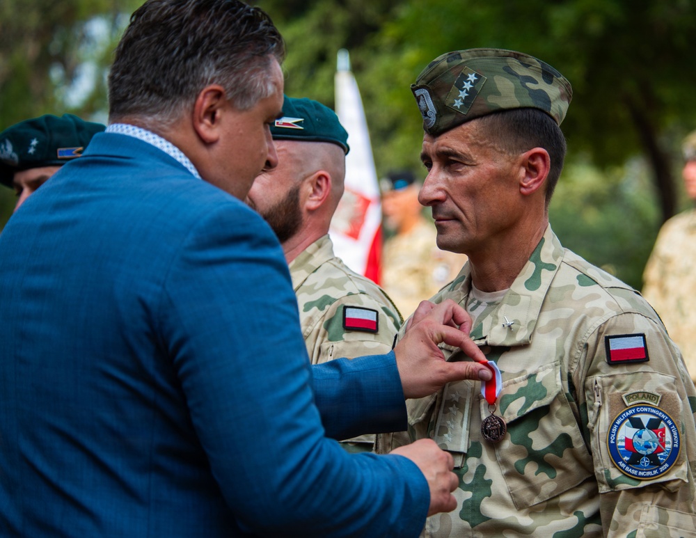 A military decoration ceremony is taking place outdoors, where a civilian in a blue suit is pinning a medal onto a Polish military officer in camouflage uniform. The officer wears a garrison cap with stars and a patch identifying 'Polish Military Contingent in Türkiye, Air Base Incirlik 2019.' Additional Polish military personnel in green berets are visible in the background.