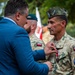 A military decoration ceremony is taking place outdoors, where a civilian in a blue suit is pinning a medal onto a Polish military officer in camouflage uniform. The officer wears a garrison cap with stars and a patch identifying 'Polish Military Contingent in Türkiye, Air Base Incirlik 2019.' Additional Polish military personnel in green berets are visible in the background.