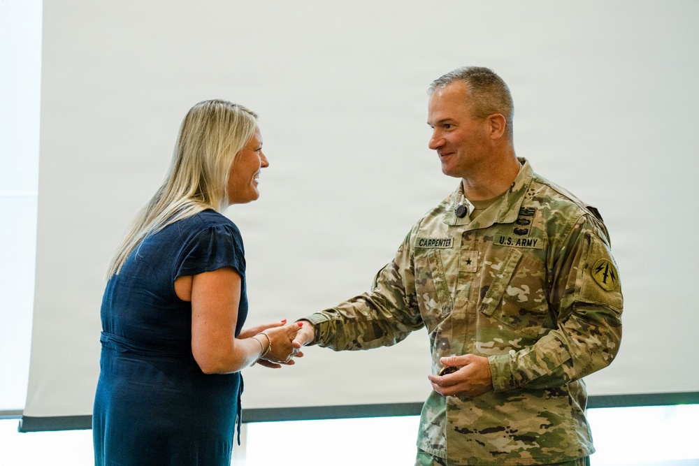 A U.S. Army officer identified by name tape as 'CARPENTER' is shaking hands with a civilian woman during what appears to be a formal recognition or award ceremony. The soldier, wearing an OCP (Operational Camouflage Pattern) uniform with a Brigadier General star insignia, is holding what appears to be a glass award or plaque. Both individuals are smiling, suggesting a positive and celebratory interaction.