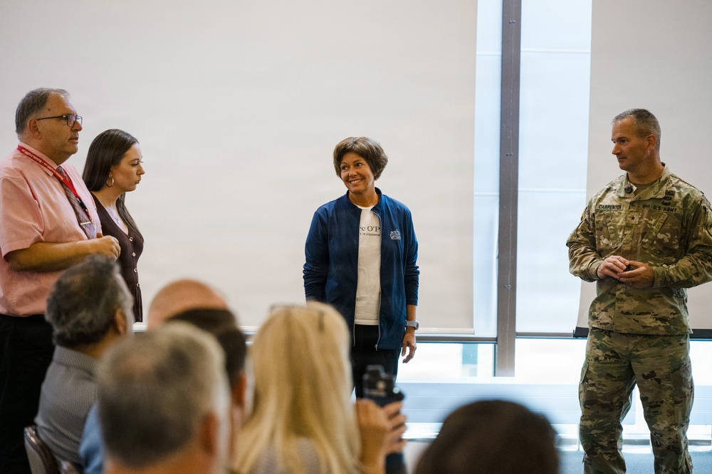 A group of four individuals are standing at the front of a meeting room, appearing to address a seated audience. One person is wearing a U.S. Army combat uniform with a name tape reading 'CARPENTER,' while the others are in civilian attire. The setting appears to be a formal or semi-formal briefing or presentation event.