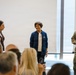 A group of four individuals are standing at the front of a meeting room, appearing to address a seated audience. One person is wearing a U.S. Army combat uniform with a name tape reading 'CARPENTER,' while the others are in civilian attire. The setting appears to be a formal or semi-formal briefing or presentation event.