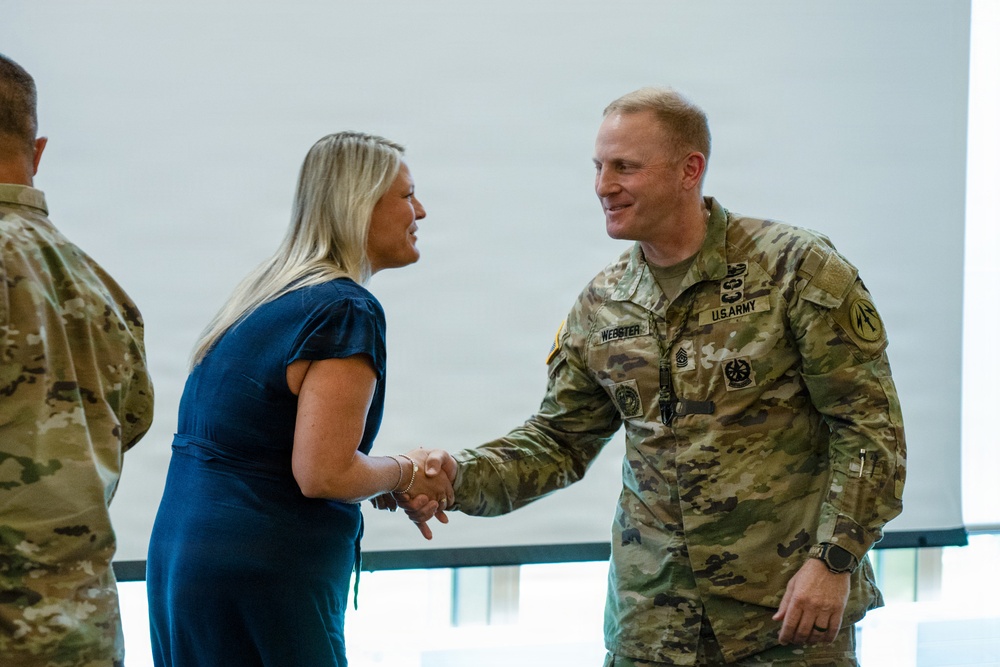 A U.S. Army soldier in OCP (Operational Camouflage Pattern) uniform is shaking hands with a civilian woman in a blue dress, both smiling. The soldier's name tape reads 'WEBSTER' and rank insignia indicates a Senior Non-Commissioned Officer. A second soldier in uniform is partially visible on the left side of the frame.