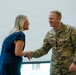 A U.S. Army soldier in OCP (Operational Camouflage Pattern) uniform is shaking hands with a civilian woman in a blue dress, both smiling. The soldier's name tape reads 'WEBSTER' and rank insignia indicates a Senior Non-Commissioned Officer. A second soldier in uniform is partially visible on the left side of the frame.