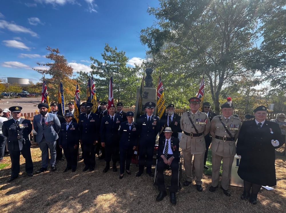 A formal military and veterans' commemoration ceremony is taking place outdoors near a memorial statue, with participants from multiple armed forces branches gathered together. The group includes British military personnel, what appear to be US Air Force members in blue dress uniforms, veterans wearing medals, and standard bearers holding British flags and Royal British Legion banners. The event appears to be a remembrance or memorial service held on a sunny autumn day.