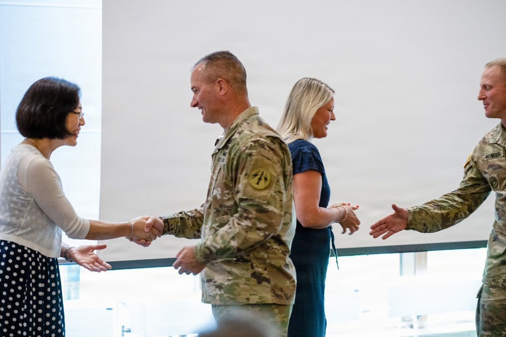A formal greeting scene showing two military personnel in camouflage uniforms shaking hands with two civilian women in what appears to be an official or ceremonial setting. The interaction appears cordial and professional, with all four individuals smiling. The setting appears to be an indoor facility with large windows in the background.
