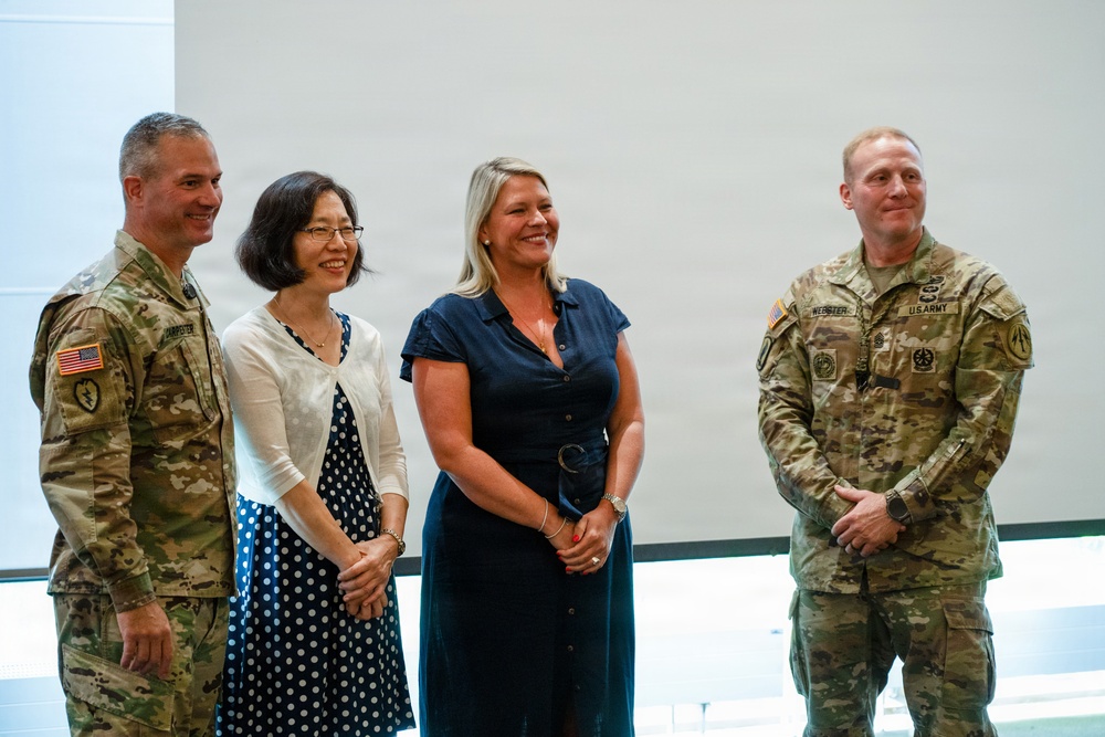 Four individuals pose together in what appears to be an indoor military or official setting. Two are wearing U.S. Army combat uniforms (OCP pattern) and two are in civilian attire. The group appears to be at a formal or ceremonial event, smiling for the photo.