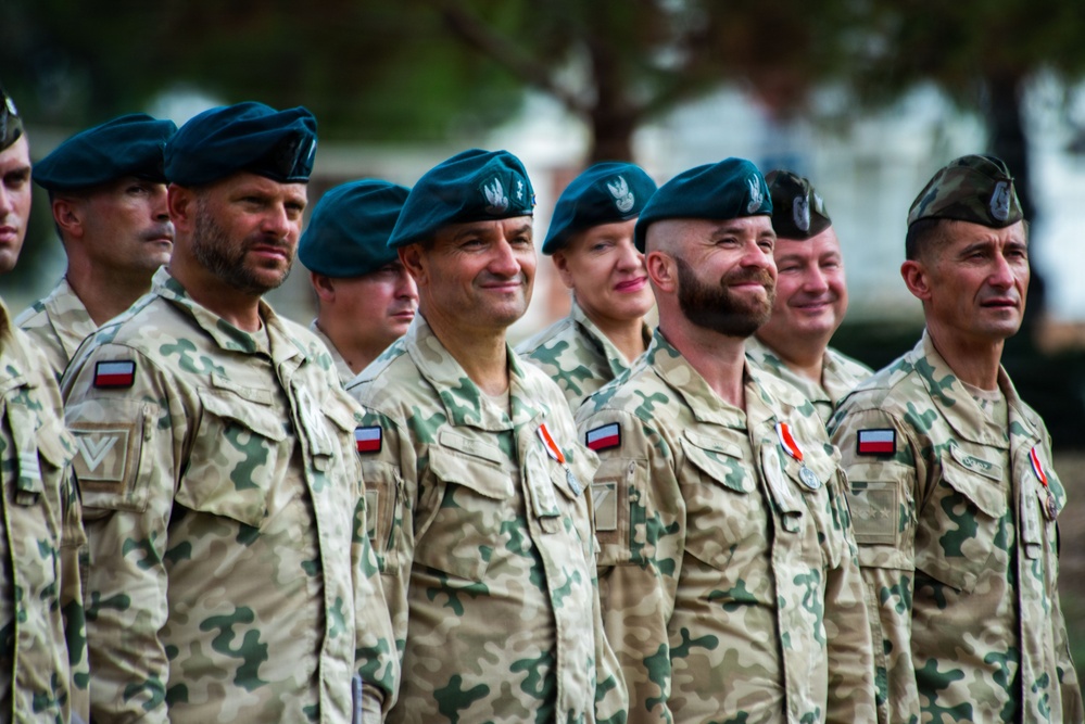 A group of Polish military personnel standing in formation during what appears to be a formal ceremony or parade. The soldiers are wearing Polish Armed Forces desert camouflage uniforms with Polish flag patches and teal/green berets bearing the Polish eagle insignia. Several soldiers have medals or ribbons pinned to their chests, indicating a decorations or awards ceremony.