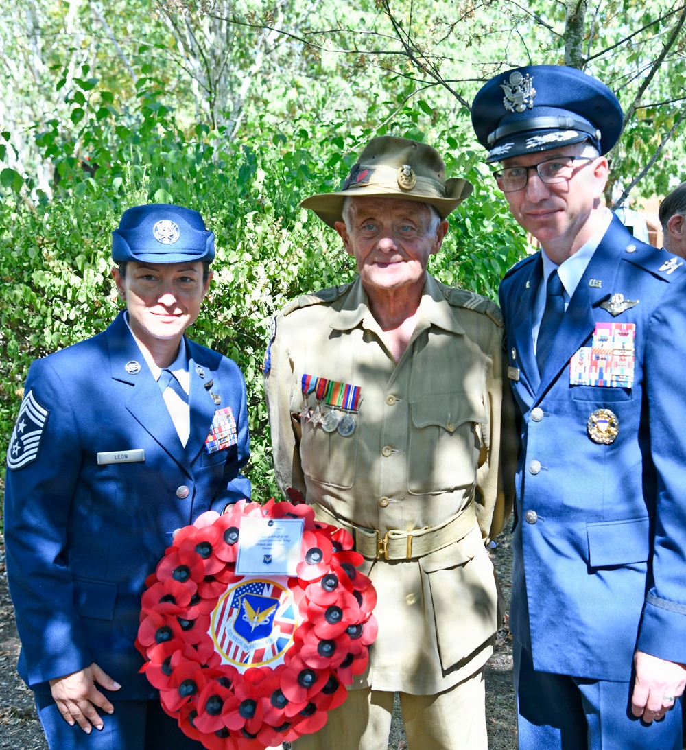 Three military personnel pose together outdoors at what appears to be a memorial ceremony. Two individuals wear US Air Force dress blue uniforms while the central figure wears an Australian Army khaki uniform with a slouch hat. They are holding a red poppy wreath bearing a combined US-UK/Allied insignia.