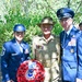 Three military personnel pose together outdoors at what appears to be a memorial ceremony. Two individuals wear US Air Force dress blue uniforms while the central figure wears an Australian Army khaki uniform with a slouch hat. They are holding a red poppy wreath bearing a combined US-UK/Allied insignia.
