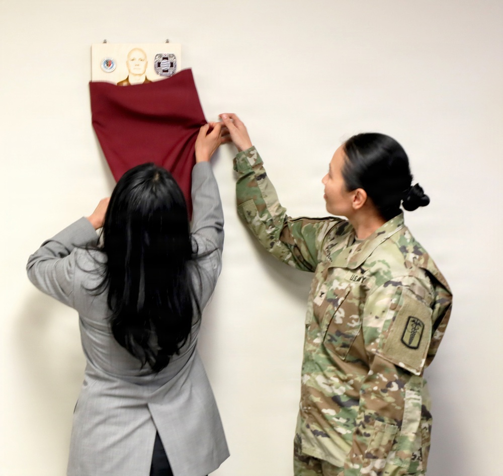 Two women are unveiling a framed display mounted on a white wall by pulling down a dark red/maroon cloth covering. One woman is in civilian attire (gray blazer) and the other is wearing a U.S. Army OCP (Operational Camouflage Pattern) uniform with a medical corps insignia patch visible. The display appears to contain military unit insignia/patches and what appears to be a portrait or bust image.