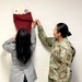 Two women are unveiling a framed display mounted on a white wall by pulling down a dark red/maroon cloth covering. One woman is in civilian attire (gray blazer) and the other is wearing a U.S. Army OCP (Operational Camouflage Pattern) uniform with a medical corps insignia patch visible. The display appears to contain military unit insignia/patches and what appears to be a portrait or bust image.