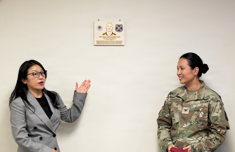 Two women stand on either side of a memorial plaque mounted on a white wall. One woman is in civilian attire (gray blazer) gesturing toward the plaque, while the other is a U.S. Army Colonel in military uniform (OCP camouflage). The plaque appears to be a dedication for the 'James Brian Connor Conference Room.'
