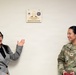 Two women stand on either side of a memorial plaque mounted on a white wall. One woman is in civilian attire (gray blazer) gesturing toward the plaque, while the other is a U.S. Army Colonel in military uniform (OCP camouflage). The plaque appears to be a dedication for the 'James Brian Connor Conference Room.'