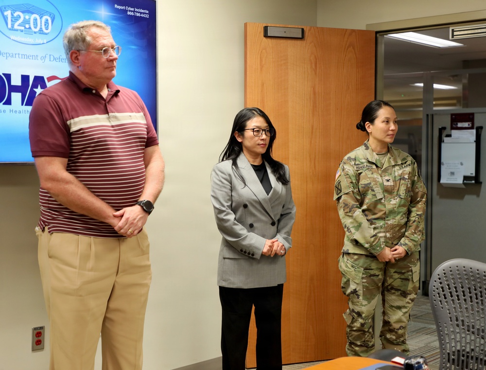 Three individuals stand in what appears to be a Department of Defense Health Agency (DHA) facility or briefing room. The group includes a civilian man in casual attire, a woman in professional business attire, and a U.S. Army soldier in Army Combat Uniform (ACU/OCP). They appear to be participating in an official meeting or presentation.