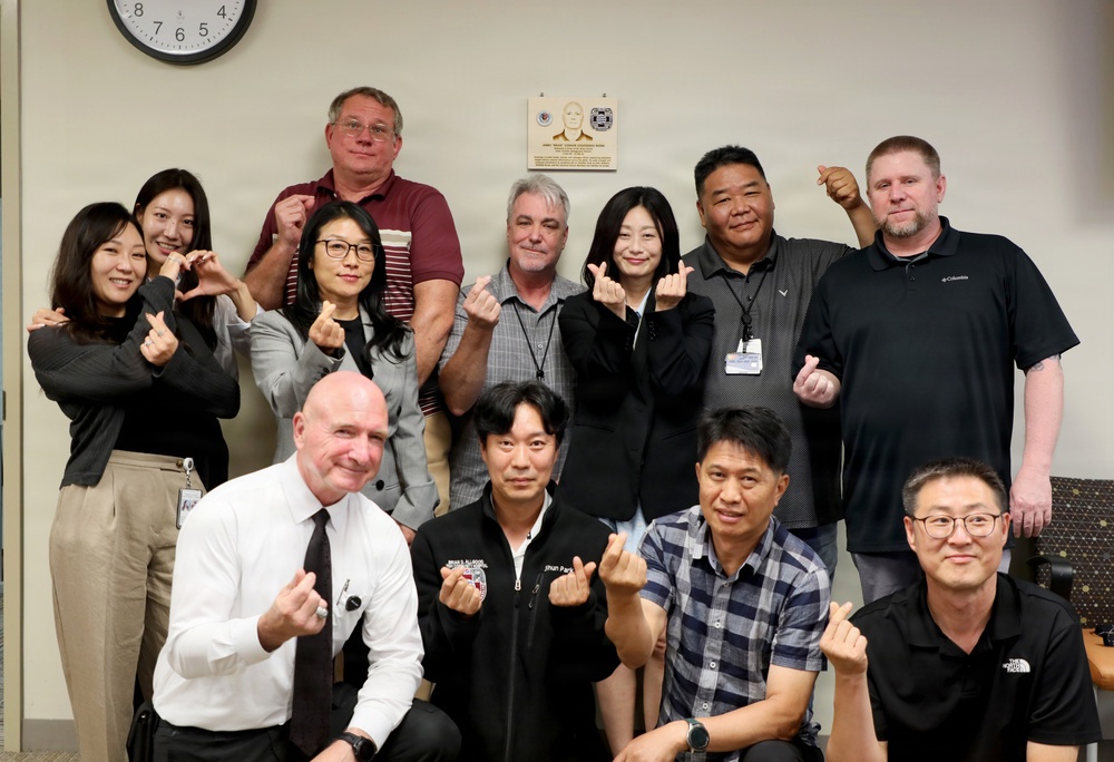 A group of approximately 12 people pose together in what appears to be an office or conference room setting. Most participants are making the Korean 'finger heart' gesture, suggesting a culturally mixed group likely celebrating or commemorating an event. A memorial or recognition plaque is visible on the wall behind them.