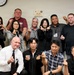 A group of approximately 12 people pose together in what appears to be an office or conference room setting. Most participants are making the Korean 'finger heart' gesture, suggesting a culturally mixed group likely celebrating or commemorating an event. A memorial or recognition plaque is visible on the wall behind them.