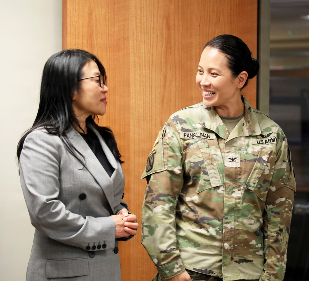 Two women are engaged in a friendly conversation. One is wearing civilian business attire (gray blazer) and the other is in U.S. Army OCP (Operational Camouflage Pattern) uniform with the name tape 'PANGELINAN' and a colonel's eagle insignia visible. The interaction appears cordial and positive, taking place indoors against a wood-paneled wall.